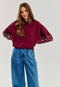 Bordowy sweter z kwiatowymi rękawami, o luźnym kroju i okrągłym dekolcie, w połączeniu z niebieskimi dżinsami o wysokim stanie.