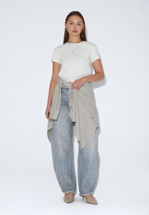 Donna con capelli castani lunghi che indossa una t-shirt bianca, jeans azzurri larghi, tacchi beige e una camicia a quadri legata in vita.
