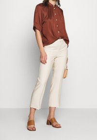 Bruine satijnen blouse met korte mouwen en knopen aan de voorkant, gecombineerd met crèmekleurige hoge flairbroeken en tan kleurige haksandalen.