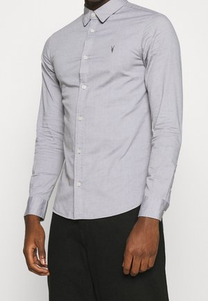 Muž nosící světle šedou košili s dlouhým rukávem a knoflíky, s malým vyšívaným logem na hrudi, v kombinaci s černými nohavicemi.