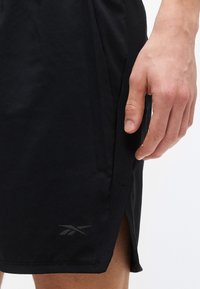 Κοντινό πλάνο του χεριού ενός ατόμου που ακουμπά δίπλα σε μαύρα σορτς με μικρό λογότυπο Reebok κοντά στο τελείωμα και πλαϊνή σχισμή.