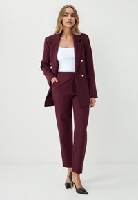 Bordeaux op maat gemaakte blazer met gouden knopen, gecombineerd met bijpassende getailleerde broek en een witte tanktop; slanke zwarte hakken maken de look af.