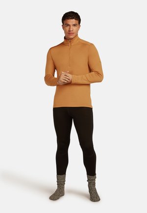 Junger Mann steht, trägt einen senfgelben Langarm-Pullover mit Viertelreißverschluss, schwarze Leggings und graue gemusterte Socken, vor einem schlichten Hintergrund.