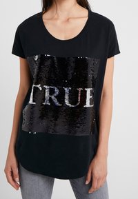Svart t-shirt med kort ärm som har ett stort paljettdesign med texten "TRUE" i svart och silver över framsidan, med rundad halsringning.