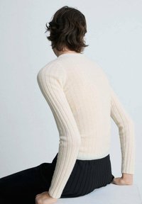 Enganliegendes, cremefarbenes, geripptes Sweatshirt mit langen Ärmeln, kombiniert mit schwarzen, strukturierten Hosen, die die Rückansicht des Outfits zeigen. Glattes Stoffgefühl.