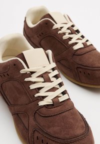 Zapatillas de ante marrón con cordones y lengüeta en crema. Las características incluyen una forma redondeada y detalles de costura sutiles. Suela de goma en marrón claro.