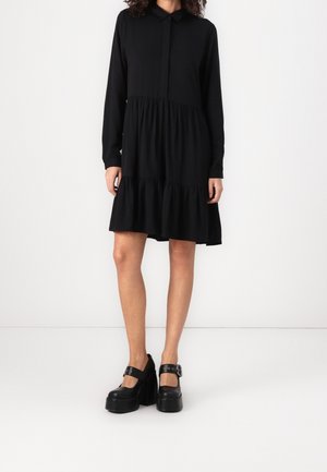 Femme porte une robe noire mi-longue à manches longues avec des volants froncés, associée à des chaussures Mary Jane à plateforme noires avec boucles latérales.