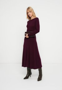 Lange paarse geribde jurk met lange mouwen, uitlopende onderkant, gecombineerd met een zwarte clutch en gedragen met kniehoge bruine laarzen.