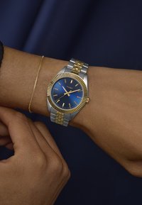 Timex-klokke med blå urskive, gull- og sølvfarget metallarmbånd, rustfritt stålhus og strukturert gullbezel; båret på håndleddet med et slankt kjedearmbånd.