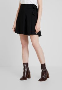 Falda negra con pliegues y detalle de cintura atada, hecha de tejido suave. Combinada con botas de tobillo burdeos brillantes con un patrón de cocodrilo.