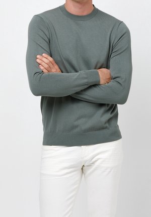 Mężczyzna z założonymi skrzyżowanymi ramionami, ubrany w oliwkowozielony sweter z długim rękawem i białe spodnie na jasnym, jednolitym tle.