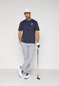 Vyriškis dėvintis baltą kepurę, tamsiai mėlyną marškinėlį, šviesiai pilkas kelnes ir golfo batelius, stovintis su golfo lazda ir apsimaušęs pirštinę priešais paprastą foną.