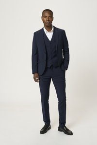 Costume trois pièces marine avec une veste à un bouton, des revers en cran et un gilet assorti. Chemise blanche en dessous, associée à des chaussures noires.