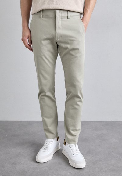 Ανοιχτό πράσινα chinos σε slim fit με λεία υφή, συνδυασμένα με άσπρα sneakers χαμηλού προφίλ. Διαθέτουν διακριτικό σχέδιο μπροστινής τσέπης.