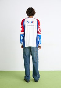 Hvid skjorte med røde, blå og flamemønstrede ærmer, med "Motorsport" logo og mønstre. Sammen med løse blå jeans.