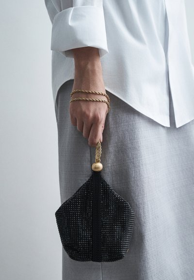 Crna perlica torbica s trokutastim oblikom, zlatnom pletenom drškom i okruglim perlicama na vrhu. Nosi se s bijelom košuljom i sivom suknjom.