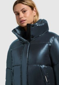 Geacă puffer de culoare albastru închis, cu un finisaj lucios, guler înalt, buzunare mari și accesorii negre. Prezintă un label vizibil al mărcii pe lateral.
