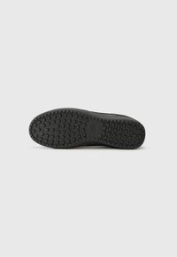 Suela de zapato de goma negra con una superficie texturizada que presenta agarraderas en forma de cuadrados y círculos para mejorar la tracción. Bordes lisos y sin marcas visibles.