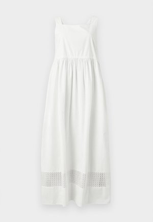 Γυναίκα που φοράει λευκό αμάνικο μάξι φόρεμα με τετράγωνο ντεκολτέ, κρατά μια μπεζ πλεκτή τσάντα και φορά καφέ σανδάλια.