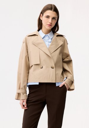 Junge Frau trägt einen beigefarbenen, gekürzten Trenchcoat über einer hellblauen Bluse mit Rüschen, kombiniert mit dunkelbraunen Hosen, steht vor einem weißen Hintergrund.