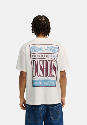 Osoba ubrana w białą koszulkę DC Shoes z dużą grafiką i napisem z tyłu, w połączeniu z niebieskimi dżinsami z naszywką z logo DC.