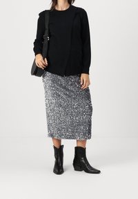 Person som bär en svart tröja, silverfärgad paljett-midi-kjol, svarta ankelstövlar och bär en svart axelväska med synlig strap-branded.