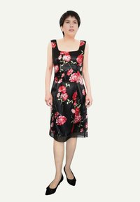 Robe noire sans manches avec un motif floral rouge vif, longueur genou, texture satinée et ourlet transparent. Portée avec des ballerines noires pointues.