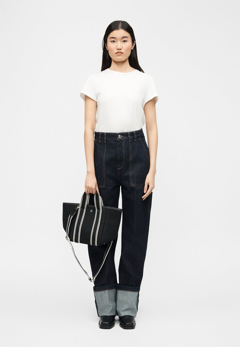 Γυναίκα με μακριά σκούρα μαλλιά που φοράει λευκό μπλουζάκι, σκούρο τζιν με γυρισμένα μπατζάκια, μαύρα παπούτσια, κρατώντας μια μαύρη τσάντα ώμου με λευκές κάθετες ρίγες.