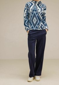 Sininen kuvioitu neule paksulla geometrisella mallilla, yhdistettynä tummansinihin leveälahkeisiin housuihin, joissa on sivunappikoristeet ja valkoiset tennarit.