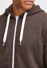 Brun zip-up hoodie med et tekstureret stof, hvide snoretræk og en høj hals. Har en frontlynlås og en afslappet pasform.