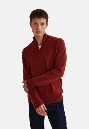 Pull en tricot rouge avec col zippé, manches longues et ourlet côtelé. Porté par-dessus une chemise blanche, associé à un jean bleu foncé.