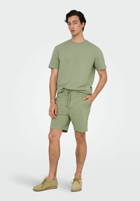 Olivengrøn tekstureret t-shirt og matchende shorts med elastisk talje. Skoene er lyse beige med snørebånd. Enkel, afslappet design.