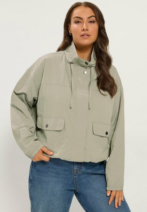 Femme portant une veste zippée vert clair avec poches à rabat et un jean bleu, debout avec une main dans la poche devant un fond uni.
