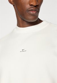 Hvid bomulds-sweatshirt med rund halsudskæring, der har et lille sort Nike-logo og teksten "NIKE STANDARD ISSUE" på brystet.