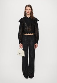 Black lace cropped blouse with ruffled shoulders, paired with black pinstriped wide-leg trousers. Model holds a small white handbag.
