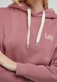 Sweat à capuche en coton rose avec cordon de serrage aux accents beiges et embouts argentés, logo brodé sur la poitrine, poignets et ourlet côtelés.
