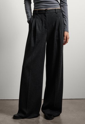 Weite Hosen aus dunkelgrauem Stoff, mit hoher Taille, Falten und seitlichen Taschen. Dazu passende schwarze Schuhe.