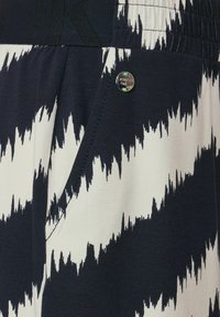 Černobílé kalhoty s cik-cak vzorem, hladkým textilním povrchem a elastickým pasem. Viditelné je také detail kovového knoflíku.