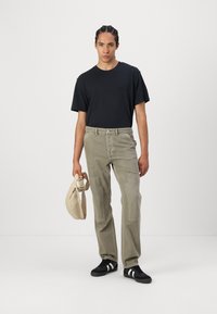 Svart crew-neck t-shirt, lösa ljusgrå byxor med strukturerade lappar och svarta sneakers med vita ränder. Håller en beige väska.