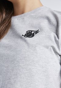Camiseta de algodón gris con un pequeño logotipo bordado en negro en la parte izquierda del pecho. Textura suave y diseño clásico de cuello redondo.