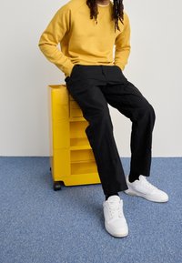 Pantaloni cargo neri e una felpa gialla con colletto a girocollo, abbinate a sneakers bianche. Sullo sfondo si vede un tappeto blu e un'unità di stoccaggio gialla.