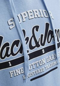 Svijetloplava dukserica s crno-bijelim grafičkim ispisom, s ključnim detaljima "SUPERIOR" i "Black&Jons". Mekani pamučni materijal.
