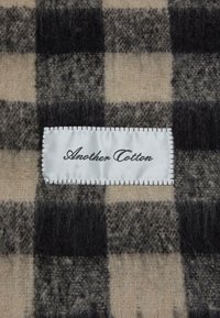 Rutig tekstil i beige og sorte nuancer med en blød, fuzzy tekstur. Et hvidt label med teksten "Another Cotton" i sort syning.