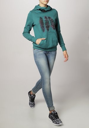 Persoon lopend met een teal hoodie met veerontwerp, licht vervaagde jeans en zwarte Adidas sneakers met patroon.