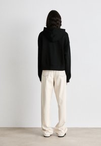 Haut à capuche noir court avec une capuche doublée, associé à un pantalon ample de couleur crème présentant des coutures visibles sur les poches arrière.