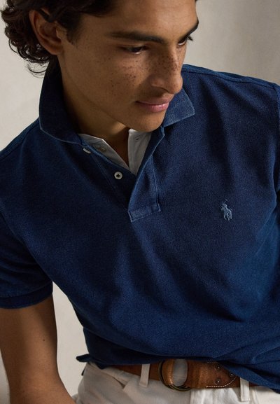 Young man with curly hair wearing a navy blue polo shirt and white pants with a brown belt, looking downward and to the side.