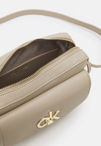 Borsa a tracolla in pelle beige di Calvin Klein con zip dorata e logo "CK" dorato sul fronte, aperta per mostrare la fodera interna con marchio.
