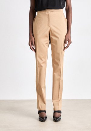 Pantaloni in tinta unita color cammello con vita alta, pieghe frontali e tasche laterali, abbinati a décolletés nere con punta a punta e cinturini alla caviglia.