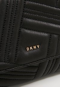 Surface en cuir matelassé noir avec lettres dorées de la marque DKNY embossées, montrant des coutures détaillées et la texture.