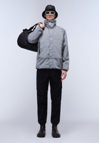 Gray insulated jacket with high collar, paired with black pants and black shoes. Model carrying a black duffel bag, wearing white sunglasses and a black bucket hat.
